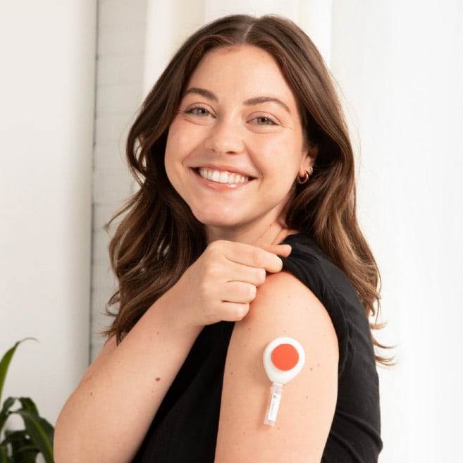 A smiling woman applies a Tasso blood lancet device to her upper arm for a simple, at-home capillary blood collection.